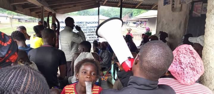 Community members gathered under a shelter at a farmers' organization meeting, with a facilitator addressing the crowd by megaphone